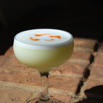 [Image shows a pale yellowish-green cocktail in a coupe glass, topped with white foam and dashes of bitters.]